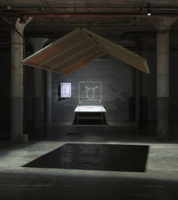 Two large stretched canvas frames hang from the ceiling of a dark space to create a roof-like structure which casts a shadow onto the cement floor with some wall-mounted works in the distance.